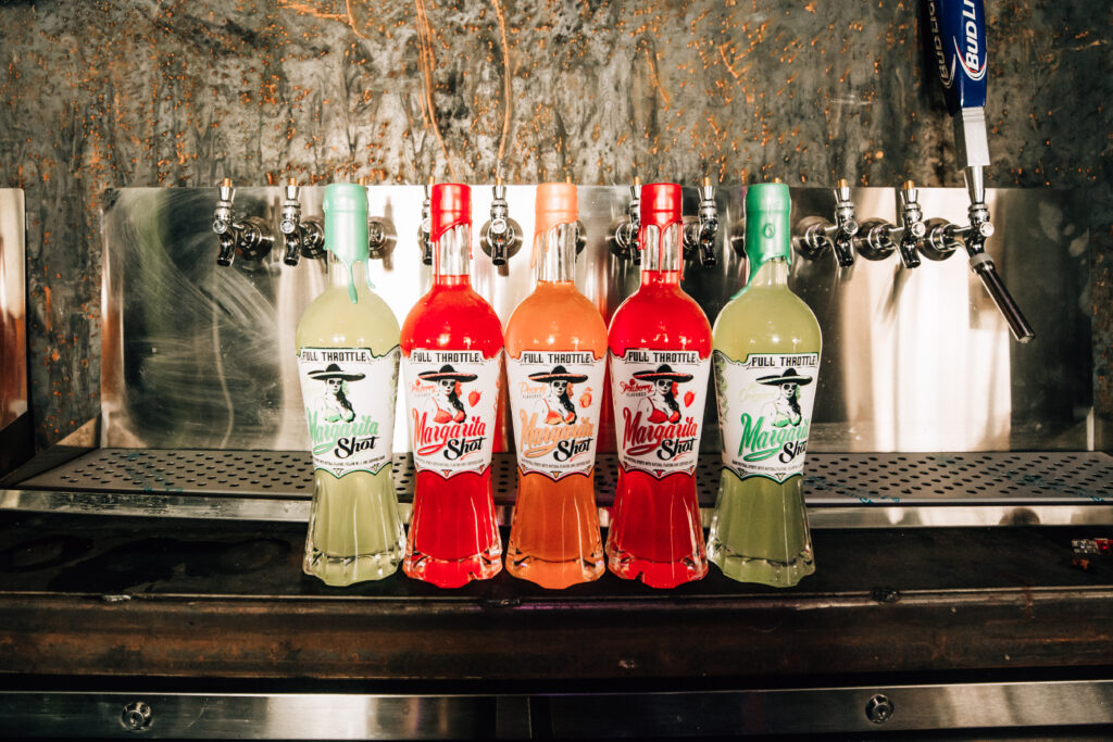 Five bottles of Full Throttle Margarita Shots in Original, Strawberry, and Peach flavors lined up on a bar in front of tap handles.
