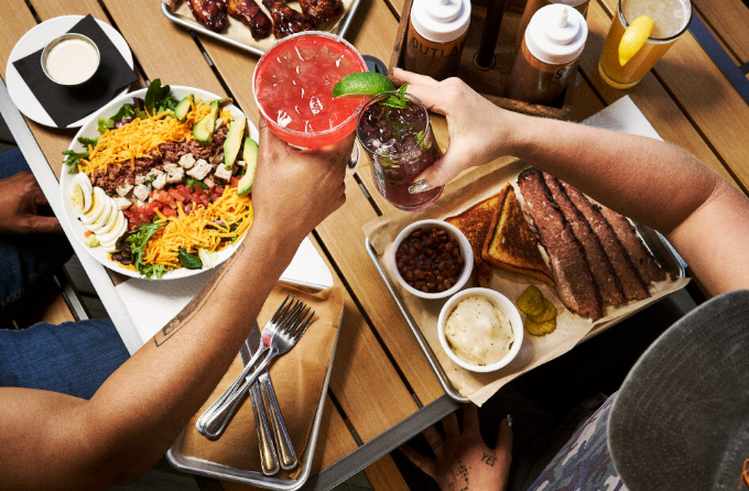 A couple toasting cocktails over a platter of smoked brisket, salad, and sides on an outdoor patio, representing a successful gathering during a fall event at Estes Park.