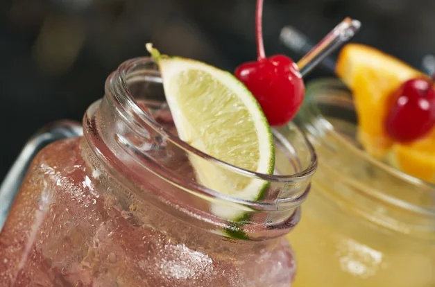 Two colorful mason jar cocktails, garnished with a lime wedge, maraschino cherries, and orange, to enjoy with a drink at Deadwood Distilling Co.